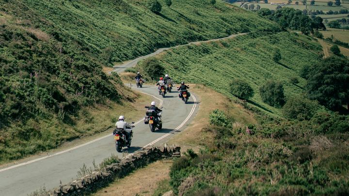Grupo de motociclistas rodando por una carretera entre colinas verdes en un día despejado